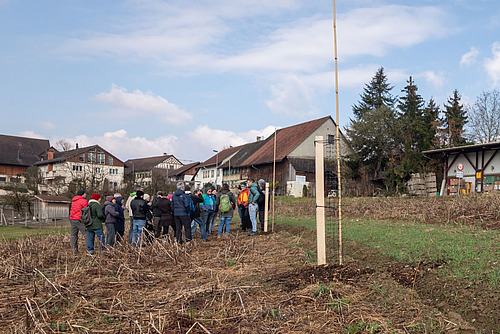 Eine Gruppe Menschen steht auf einem Acker, im Vordergrund ist ein frisch gepflanzter Baum zu sehen. Der Baum hat noch kein Laub, der Acker ist noch ohne Kultur.  